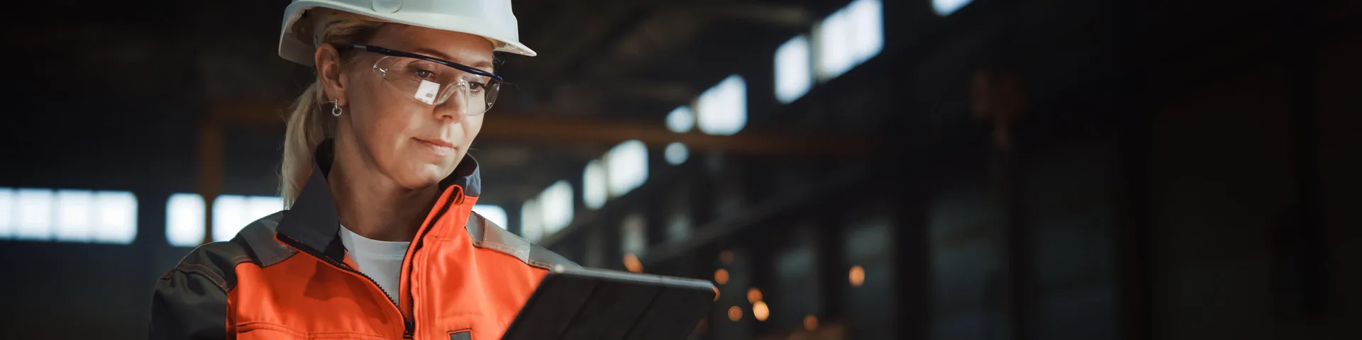 Professional Heavy Industry Engineer Worker Wearing Safety Uniform and Hard Hat Uses Tablet Computer. Serious Successful Female Industrial Specialist Standing in a Metal Manufacture Warehouse.