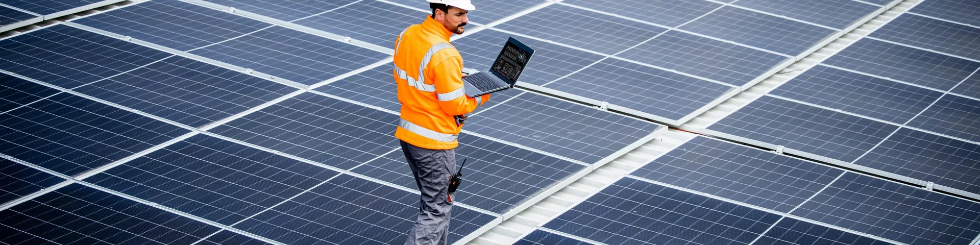 Renewable energy engineer walking through large solar power plant and checking electricity production on his laptop computer. Sustainable energy source and eco friendly technologies.