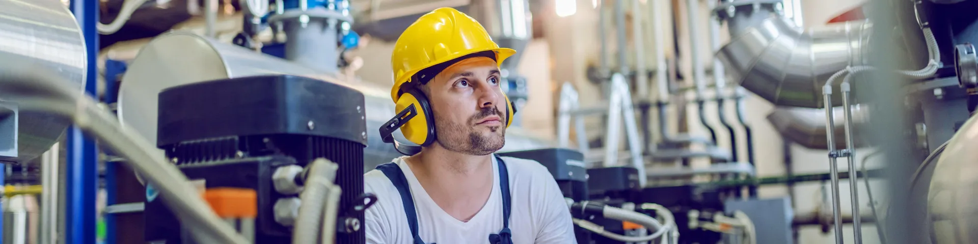 Serious caucasian handsome worker in overall, with hardhat and antiphons crouching and using tablet. Factory interior.